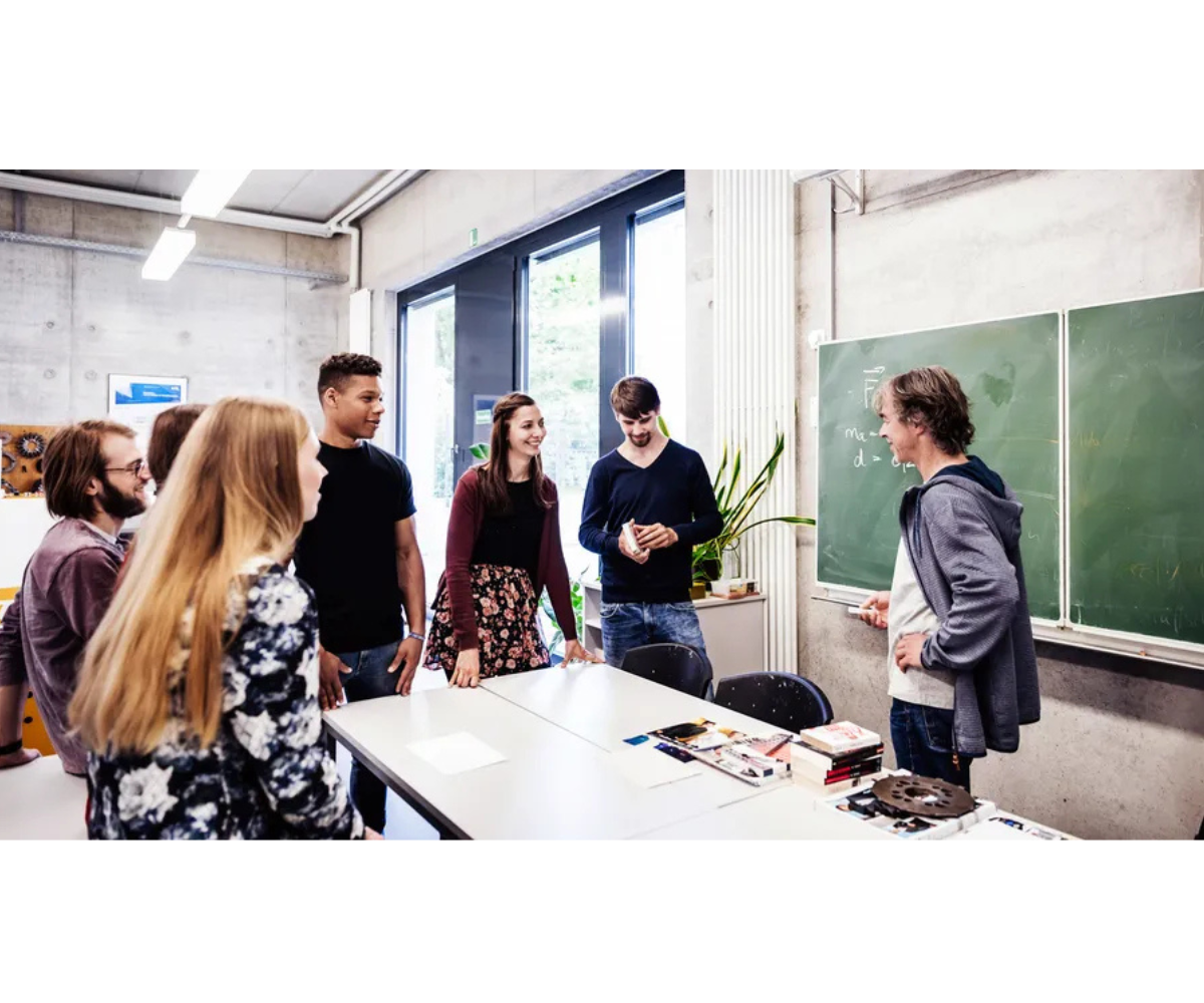 A group of students gathers around a classroom table, engaging in an interactive discussion with their teacher near a chalkboard.