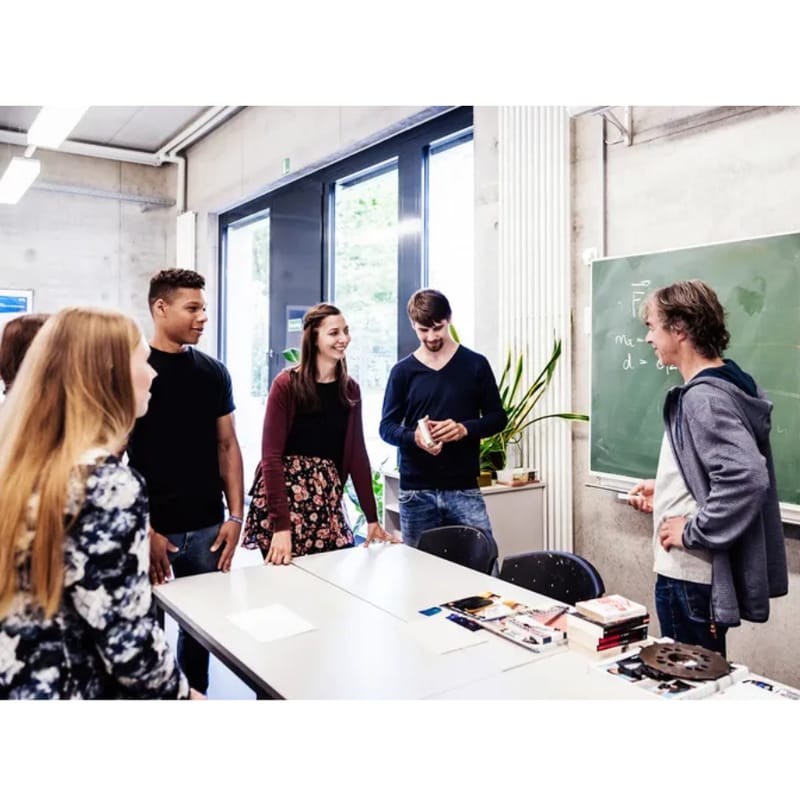 A group of students gathers around a classroom table, engaging in an interactive discussion with their teacher near a chalkboard.