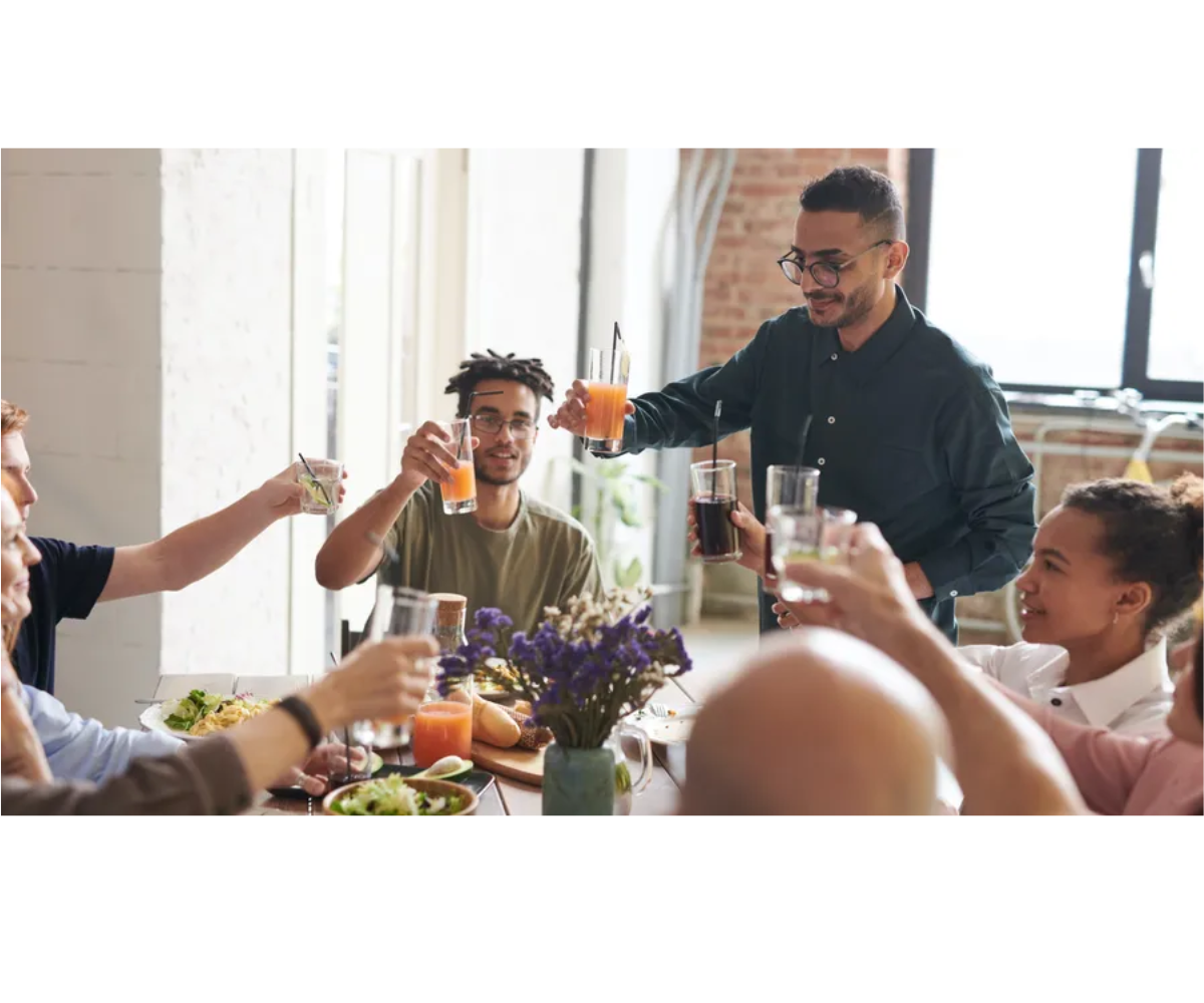 Friends gather around a table, smiling and raising drinks in a cheerful toast during a casual meal indoors.