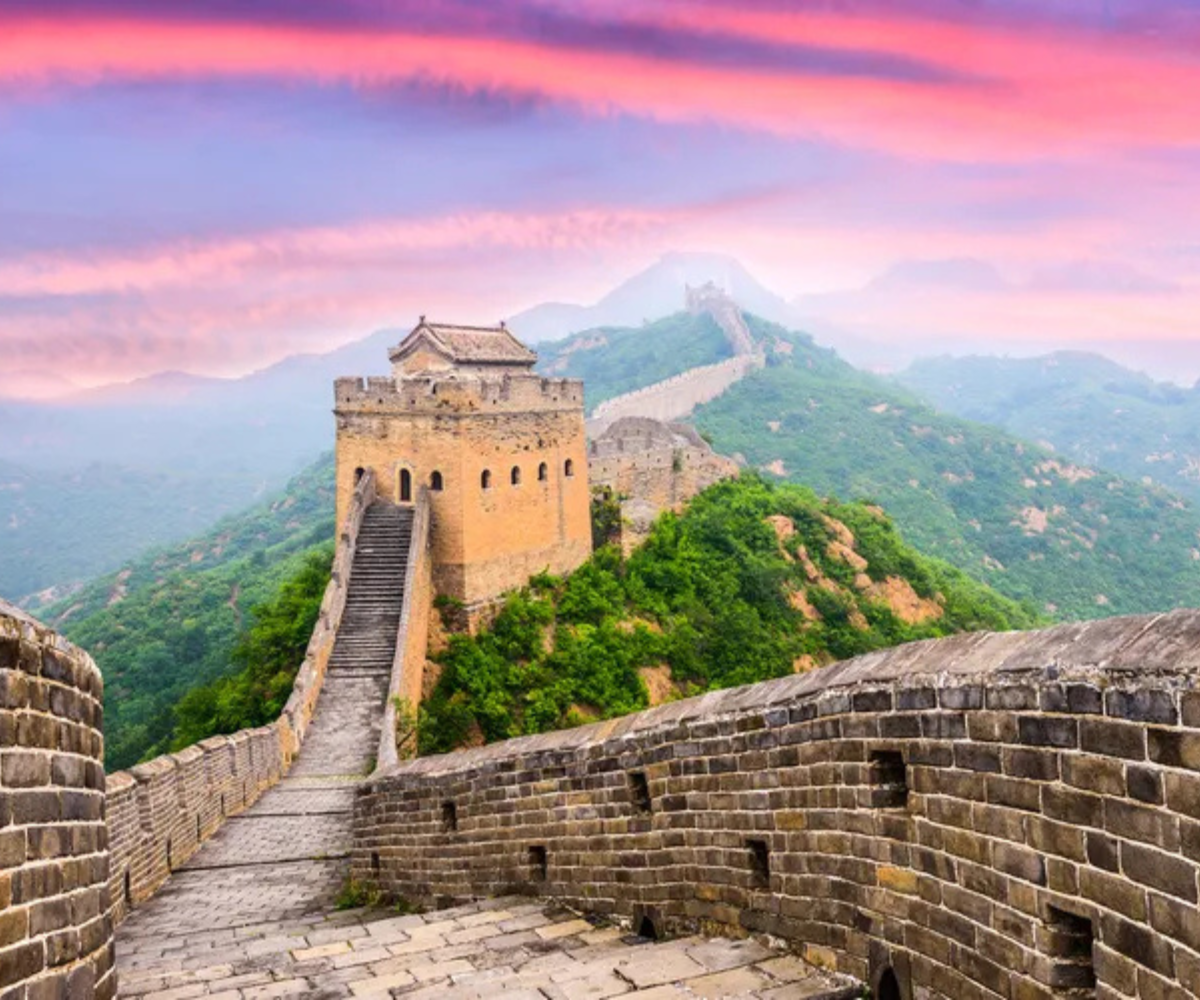 A scenic view of the Great Wall of China stretching across lush hills under a colorful pink and blue sunset sky.