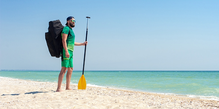 Homme à la plage avec un sac à dos pour une stand up paddle gonflable sur le dos