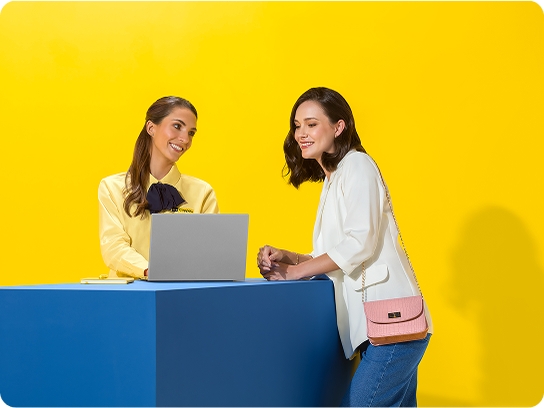 Dos mujeres platicando sobre una mesa contra un fondo amarillo brillante.