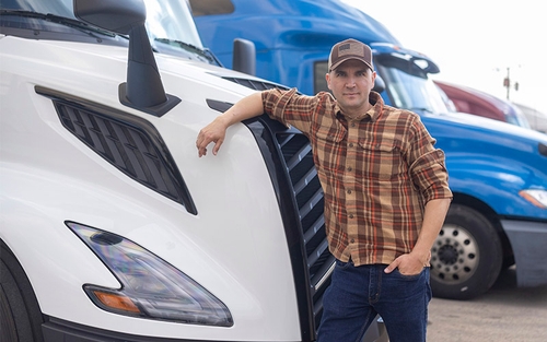 A man leaning on the front of a semi-truck wearing a plaid flannel shirt and a baseball hat with an American flag on it.