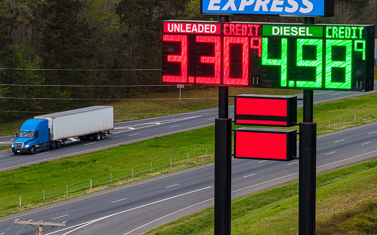 A semi-truck driving down a highway and a fuel sign with diesel prices listed at $4.98 and unleaded gasoline at $3.30.