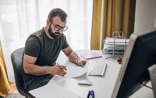 A man with glasses sitting at a desk looking down at a piece of paper and holding a pen in his hand.