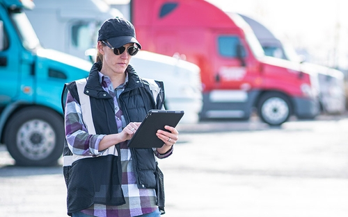 A female owner-operator standing in a yard with semi-trucks in the background, wearing a hat, sunglasses and a black vest, typing on a tablet.