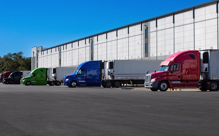 Five semi-trucks with trailers parked at a loading dock.