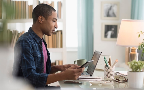 A man in a flannel shirt checks his phone while sitting at a desk that holds a laptop, coffee mug and various pens.