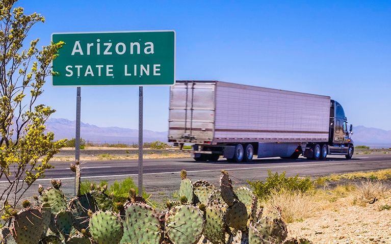 A semi-truck driving down a county highway with a green sign that says, "Arizona, state line" in the forefront.
