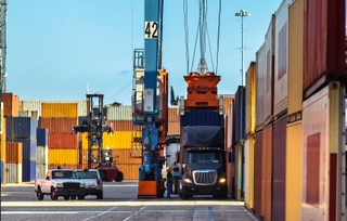 An ocean container being placed on a semi truck chassis with a crane with stacks of containers all around and in the background