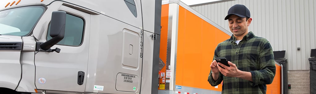 An owner-operator using his phone to look at the Schneider FreightPower app while standing outside his truck and Schneider trailer at a loading dock.