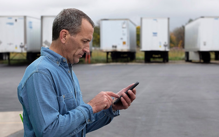 A man stands in front of a row of semi-trailers while looking at his smart phone.