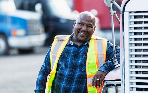 A man leaning on a semi-truck, smiling, wearing a blue plaid shirt and a yellow safety vest.