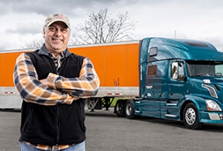 Un operador propietario con los brazos cruzados se para frente a su camion, transportando un tráiler de Schneider. 