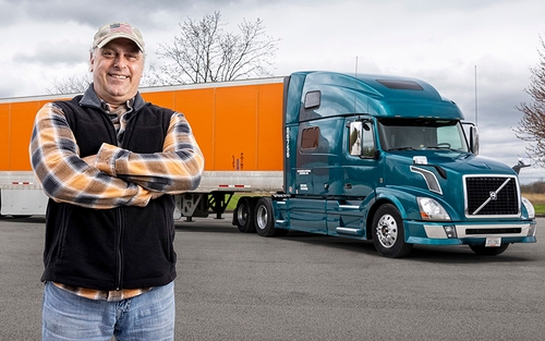 A man standing in front of a semi-trailer smiling with his arms crossed in front of him. The tractor is blue and the trailer is orange.