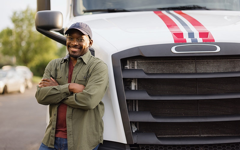 A man wearing a baseball cap leaning against a white semi-truck and smiling with his arms crossed in front of him.