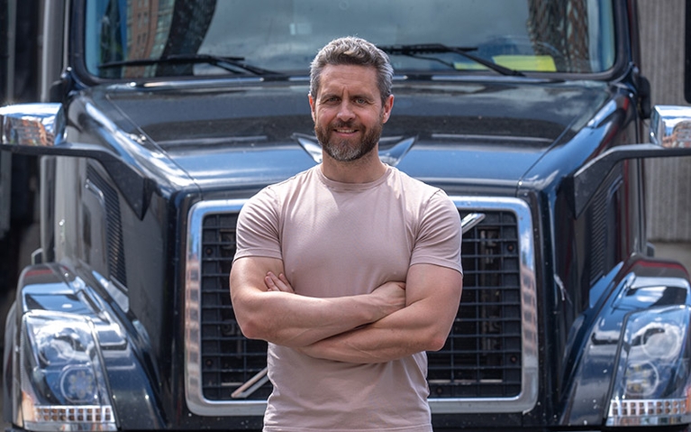 A man standing in front of a semi-truck. He's smiling and has his arms crossed in front of him.