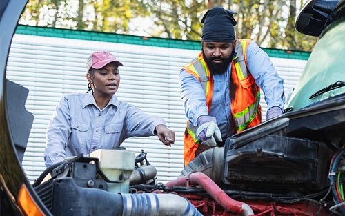 A female and male diesel technician standing outside pointing to various components under the hood of a semi-truck.