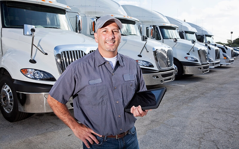 A man in a grey shirt and baseball hat stands in front of a row of white semi-trucks while holding a tablet.