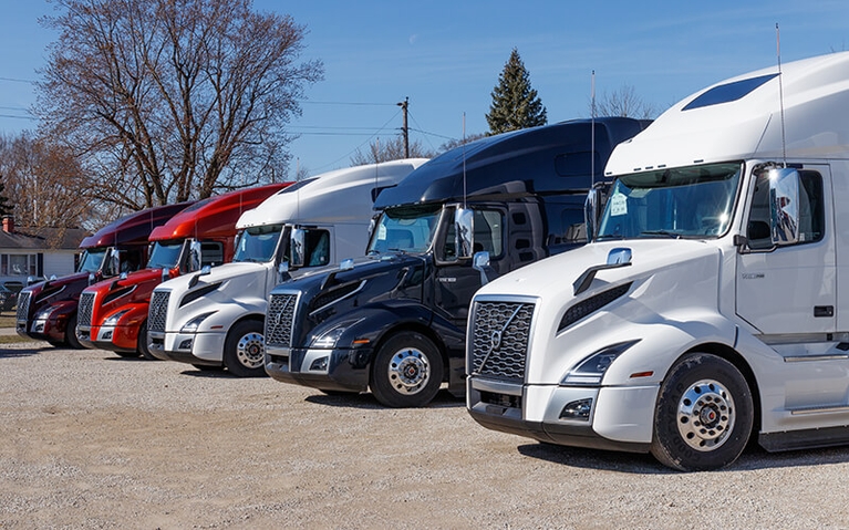 A row of five Volvo tractors, varying in color, are parked in a gravel parking lot.