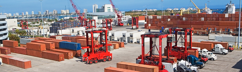 An active port with cranes, semi trucks and several rows of ocean containers stacked in the yard