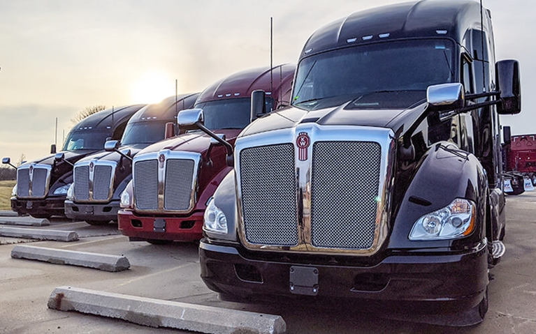 Four different colored Kenworth semi-trucks are lined up in a row in a parking lot.