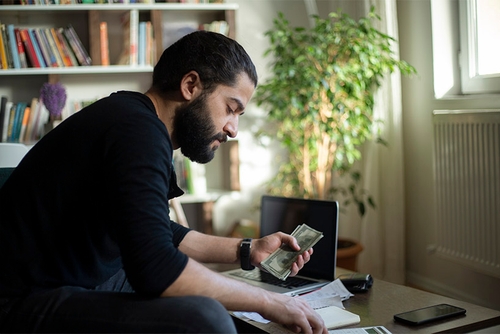 A man sitting in a living room holding a stack of cash and using a calculator.