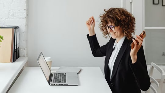 Business professional at a desk using a laptop and holding a phone while gesturing during work.