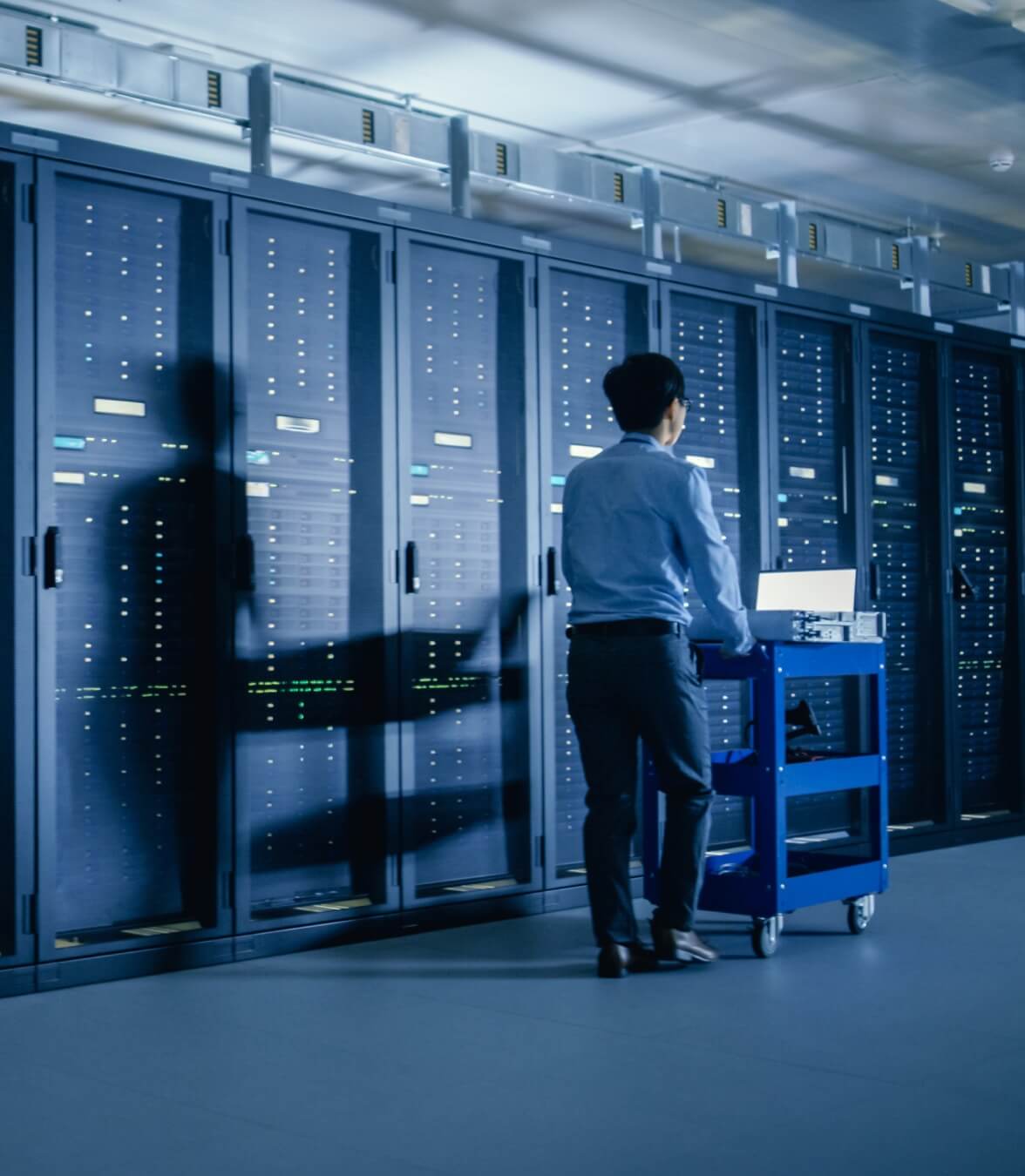 A person standing in a data center aisle while working on equipment in front of tall server racks.