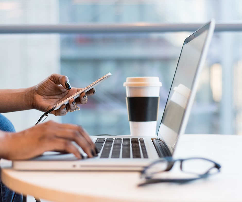 closeup of workstation with laptop, computer glasses, coffee cup and the person's hands - one on the keyboard and the other holding smart phone