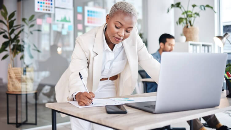 person standing and taking notes on paper from laptop