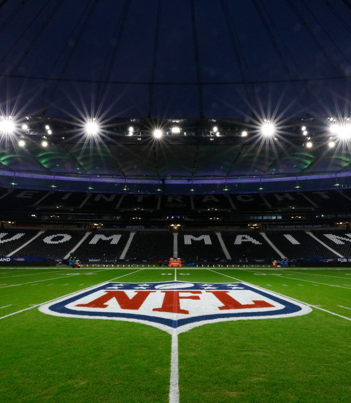 Vista noturna de um campo de futebol americano iluminado da NFL, com o logotipo da NFL no meio do campo dentro de um grande estádio.
