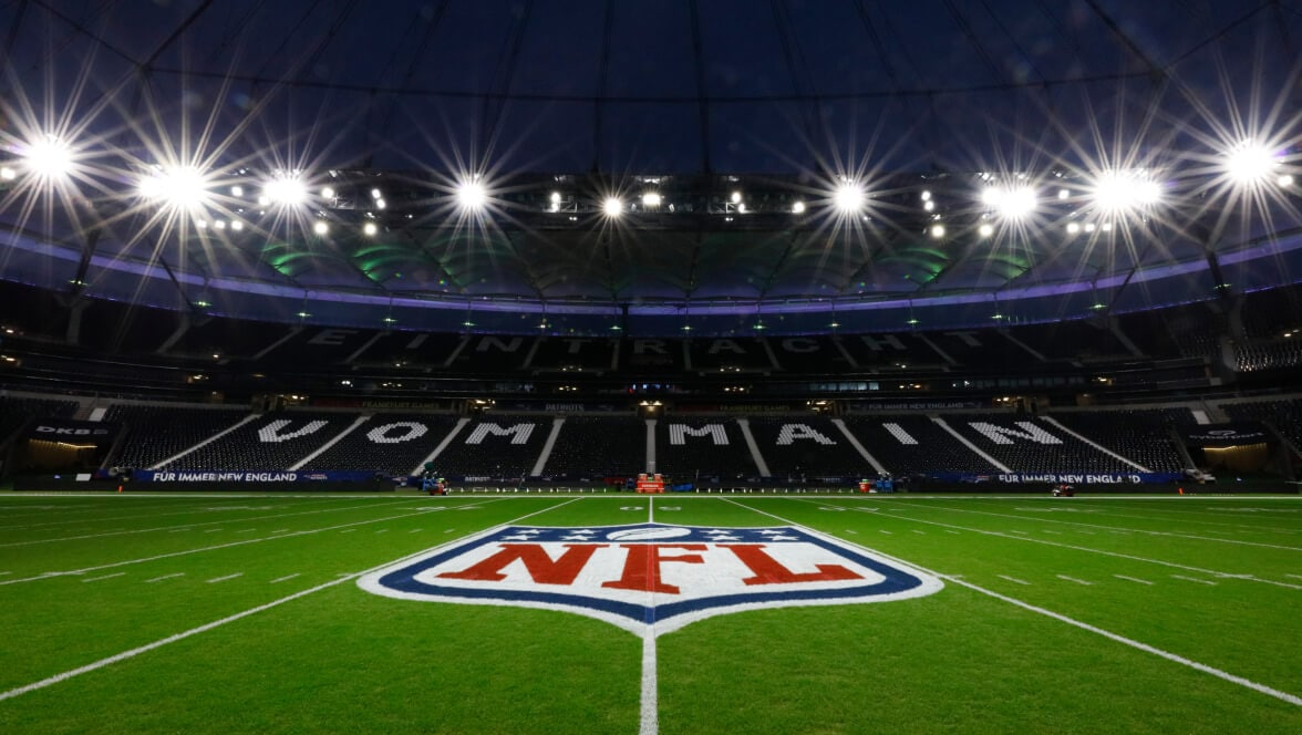 NFL field at night with the NFL shield logo at midfield inside an illuminated stadium.