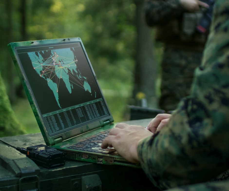 Person in camouflage using a rugged laptop with a global map and network data in an outdoor forest setting.