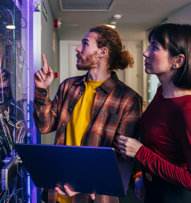 Two people collaborating in a server room while reviewing data on a laptop.