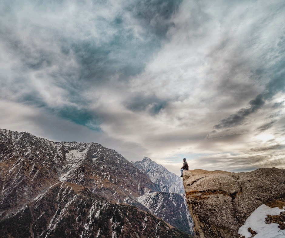 person sitting on cliff overlooking snow-capped mountains