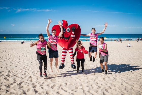 A group of people and a heart mascot jumping for joy on the beach.