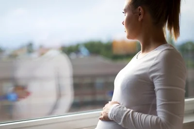Woman in white long sleeve top looking out window with one hand placed on her pregnant belly.