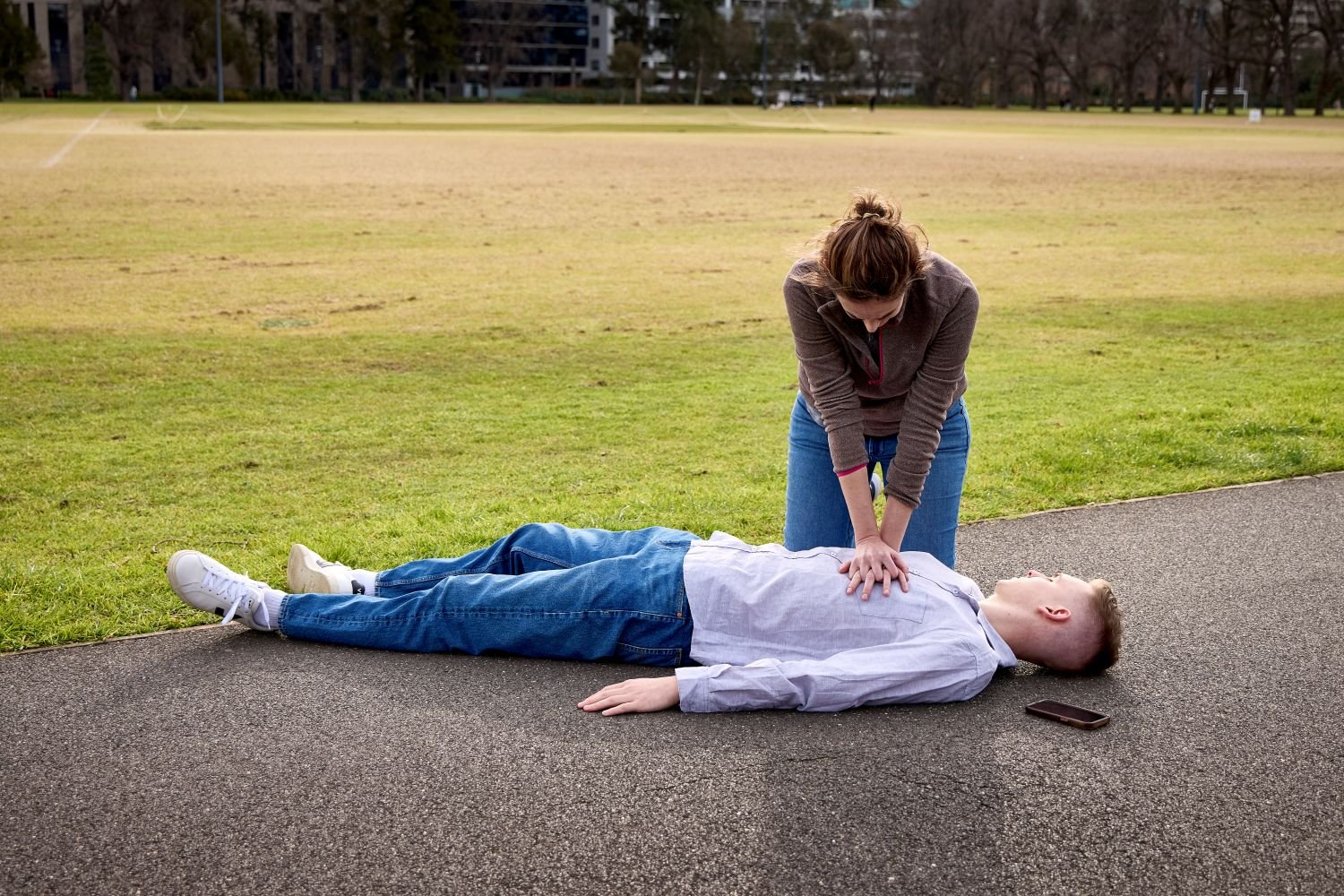 Person performing CPR on an unresponsive person lying on a path beside a sports field.