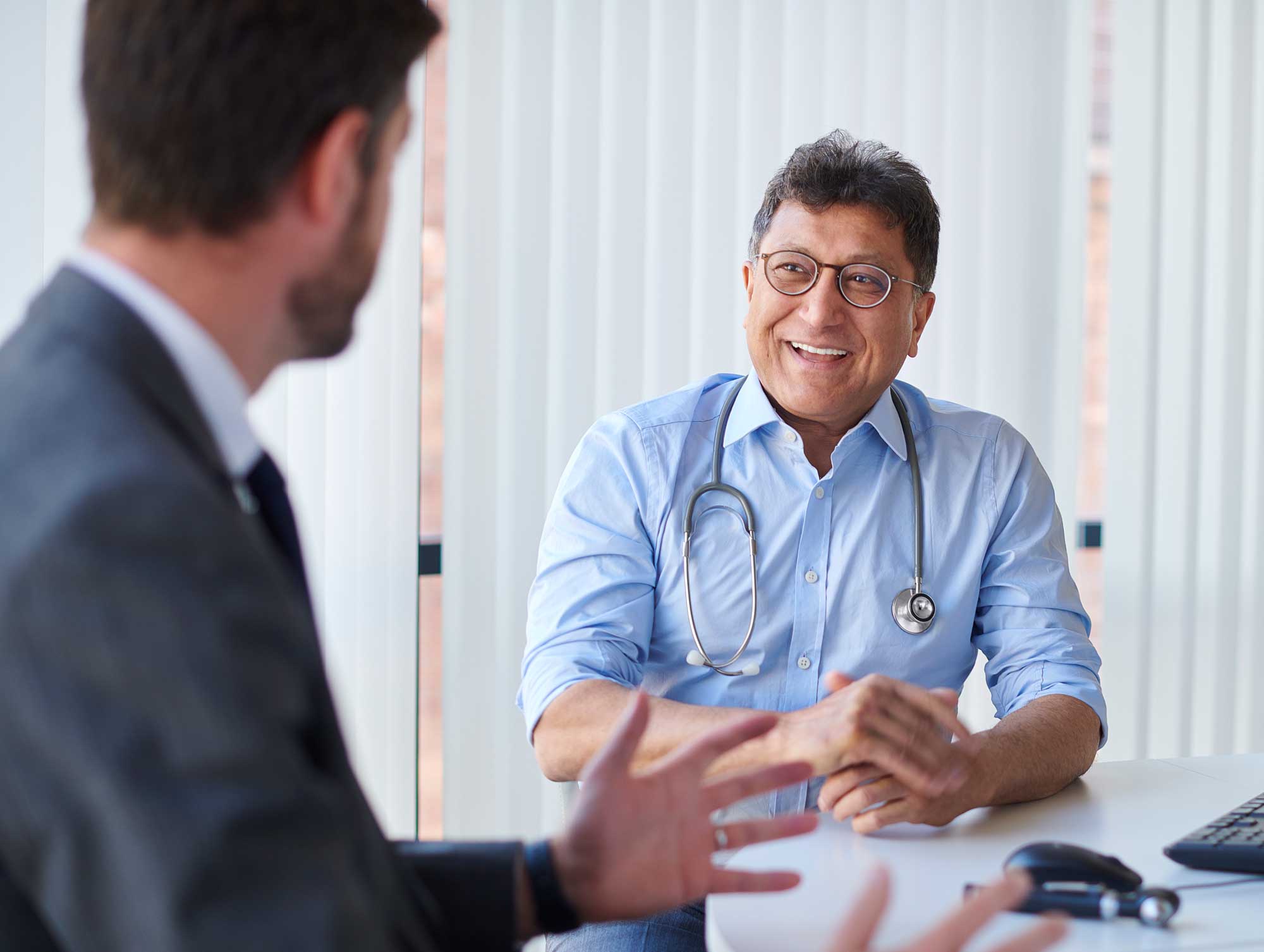 Smiling general practitioner is speaking with another man across his desk