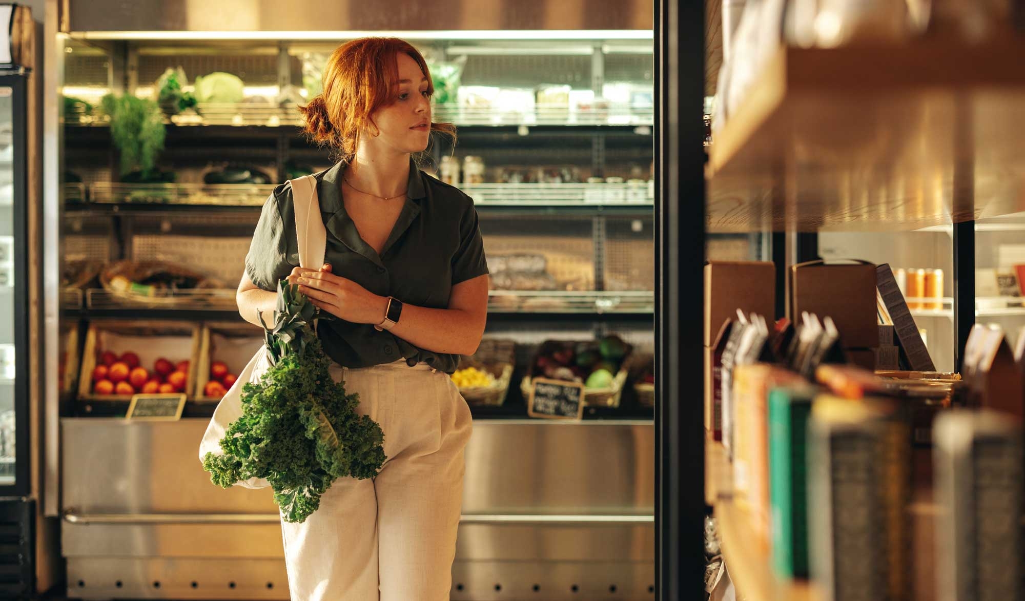 a young woman is browsing grocery store looking for fresh produce, she is carrying a bag with greens spilling out