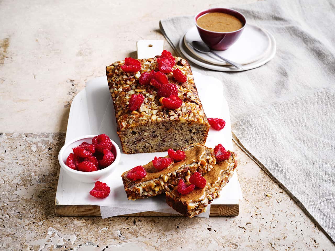 A load of Nutty Banana Loaf with a slice on a plate and a small bowl of raspberries next to it