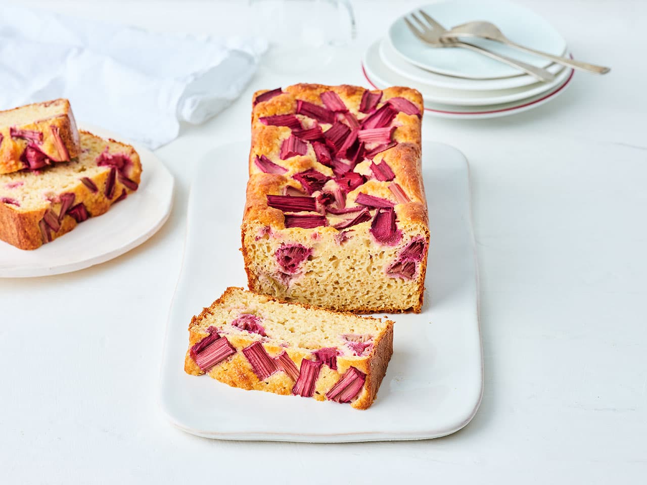 A rectangle loaf of Lemon Rhubard Yoghurt Cake, with a slice cut on a plate