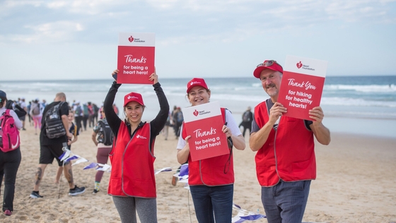 Three volunteers in red vests holding up signs on the beach, promoting the Coastrek 2024 event.