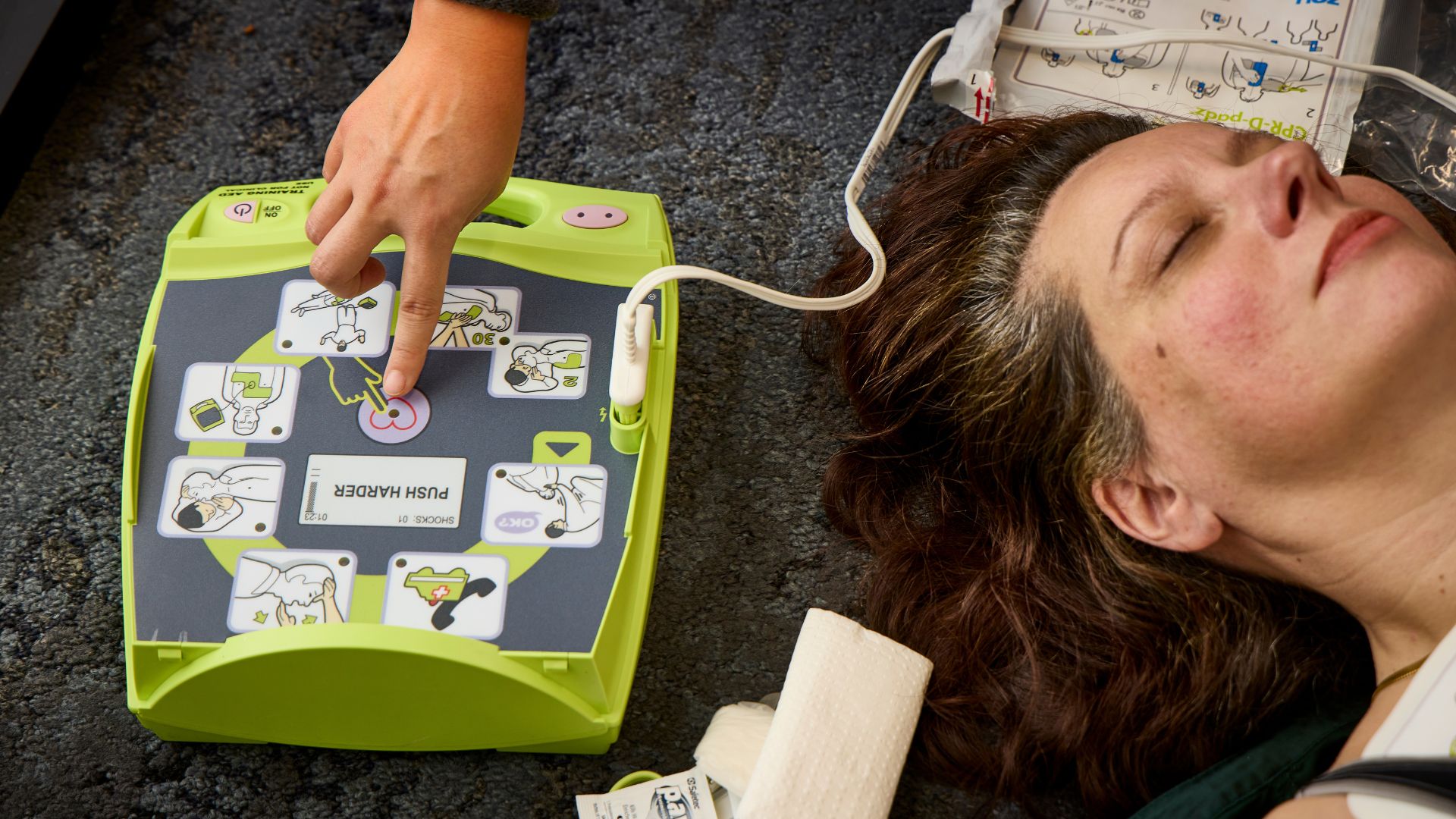 A person lies unconscious on the floor as an AED is activated nearby, with visible medical supplies and illustrated instructions on the device.