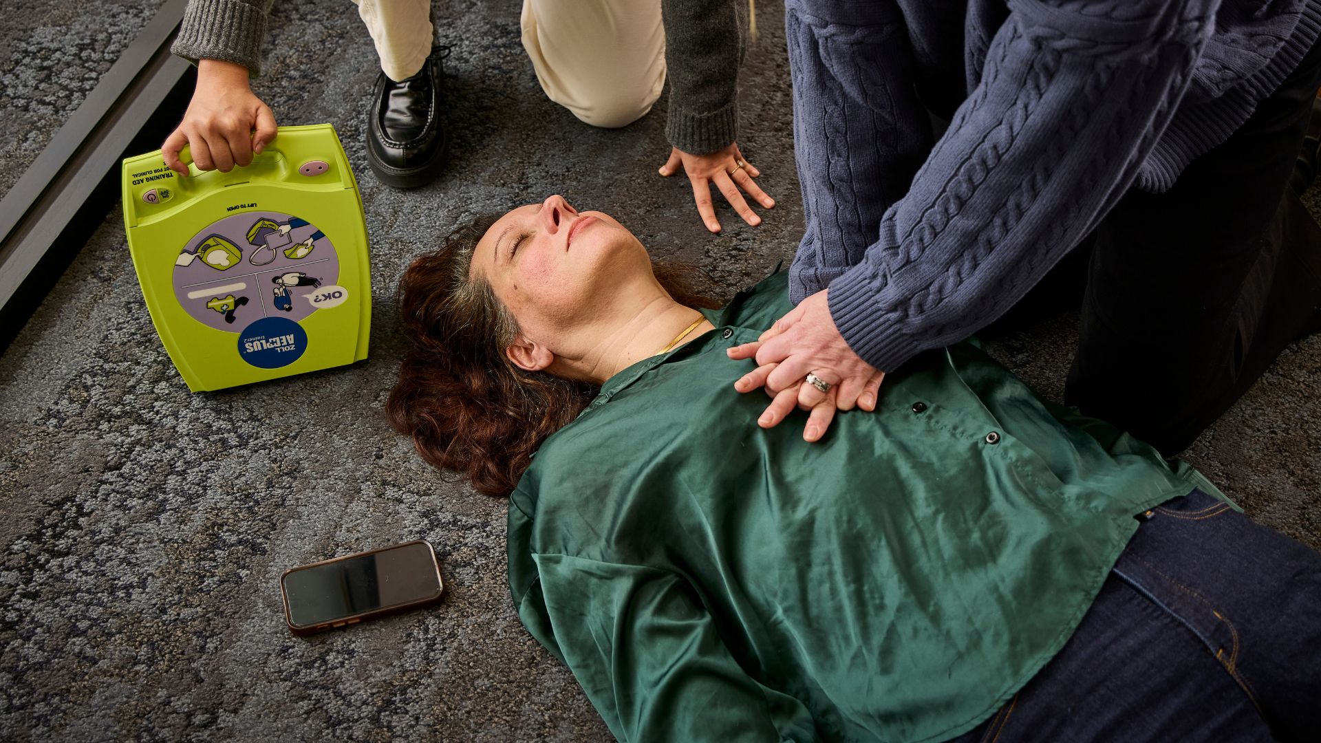 A person receives CPR on the floor while another prepares an AED; a smartphone lies nearby, capturing an urgent emergency response scene.