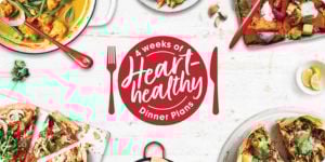 A variety of colorful and healthy dishes are arranged around a central red circle that reads '4 weeks of Heart-healthy Dinner Plans' with a red fork and knife on either side. The dishes include vegetable curry, a mixed vegetable stir-fry, roasted vegetables, two types of pizza with various toppings, and a dish with tortilla chips. There are also lemon wedges and garlic cloves scattered around the table.