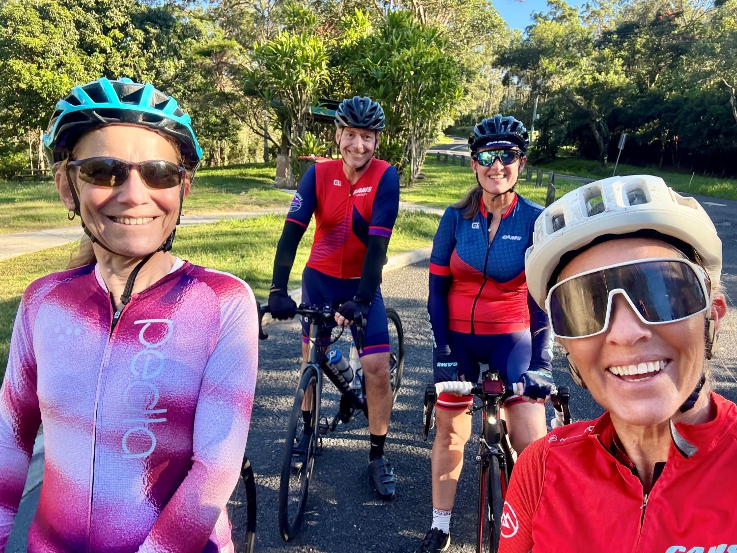 Four cyclists wearing helmets and cycling gear smile for a group selfie on a quiet, tree-lined road during a sunny ride.