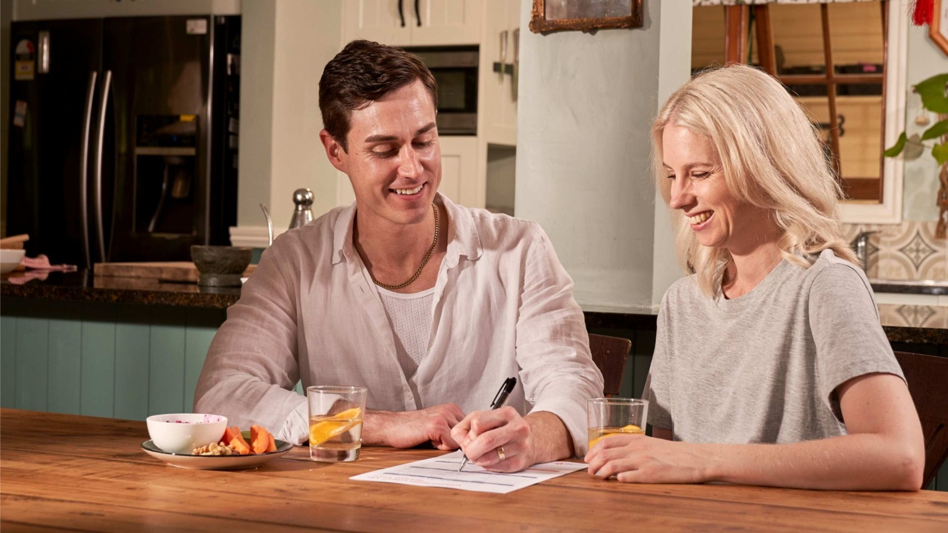 A couple sitting at a wooden table in their kitchen, writing their Will. The man, wearing a white shirt, is holding a pen and writing on a piece of paper while the woman, in a gray t-shirt, looks on. 