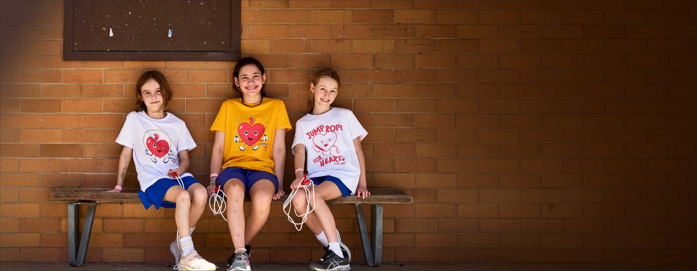 Children wearing Jump Rope for Heart t‑shirts sitting on a bench holding skipping ropes